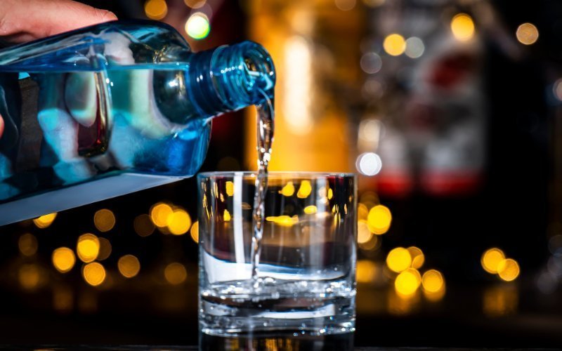 An up-close shot of a clear liquid being poured from a distinctive blue glass bottle into a clear, short tumbler glass. The stream of liquid is captured mid-pour, splashing slightly at the bottom of the glass. The background is softly blurred with warm, glowing yellow and white bokeh lights against a dark setting, creating a cozy nightlife or bar atmosphere.